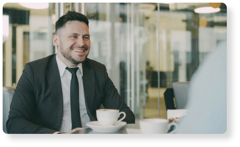 Smiling business professional in a suit and tie sitting across from a colleague during a meeting, with a coffee cup on the table.